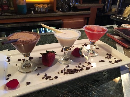 Three colorful cocktails on a white platter, garnished with strawberries and chocolate shavings.
