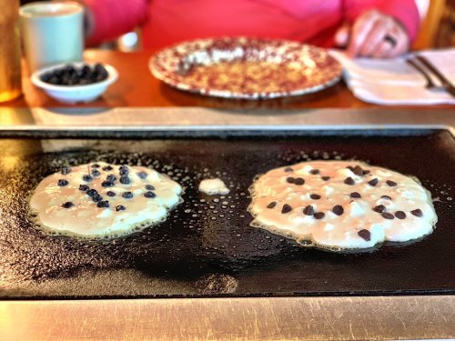 Two pancakes cooking on a griddle, topped with blueberries and chocolate chips, with a plate and coffee in the background.
