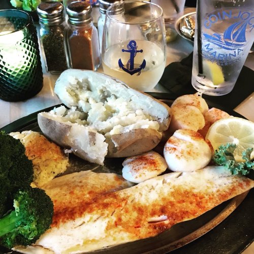 A plate of fish with scallops, baked potato, broccoli, and cornbread, accompanied by drinks and condiments.