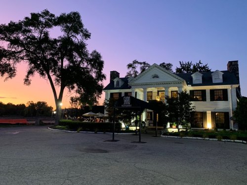 A charming white mansion with black shutters, illuminated at dusk, surrounded by trees and a colorful sunset sky.