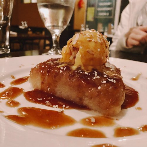 A dessert featuring a glistening cake topped with a scoop of ice cream and drizzled with sauce, garnished with sesame seeds.