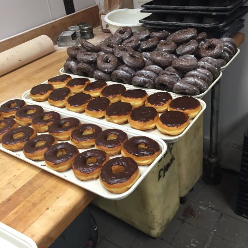 Trays of freshly made donuts, including chocolate-glazed and dark chocolate varieties, on a wooden countertop.