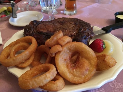 A plate featuring a large steak, onion rings, and a strawberry, set on a table with drinks in the background.