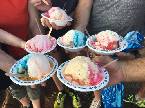 A group of hands holding colorful snow cones in paper cups, featuring vibrant flavors like blue, pink, and orange.