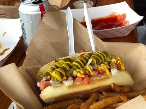 A hot dog topped with mustard, onions, and tomatoes, served with fries and a soda in a casual dining setting.