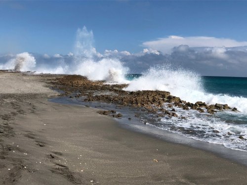 Waves crashing against rocky shore under a clear blue sky, with gentle clouds in the background.