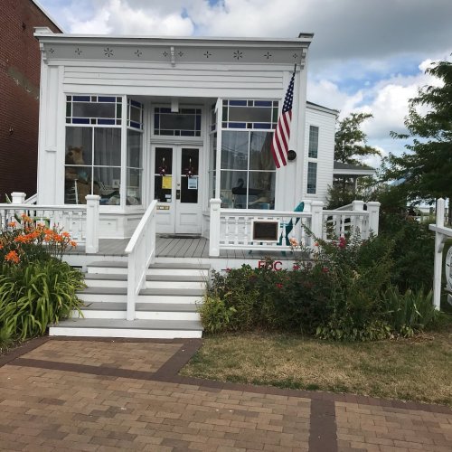 A white building with blue accents, an American flag, and colorful flowers in front, set against a cloudy sky.