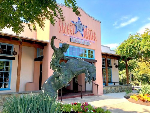 A bronze statue of a dancer in front of the Sweetwater Tavern, a colorful building with large windows and greenery.