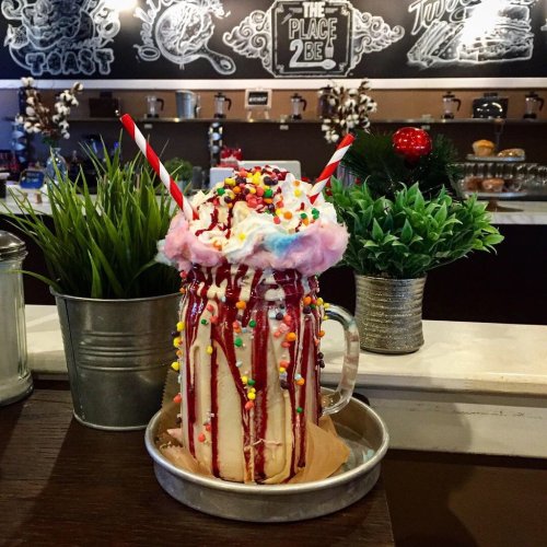 A colorful milkshake topped with whipped cream, cotton candy, sprinkles, and drizzled with syrup, served in a glass mug.