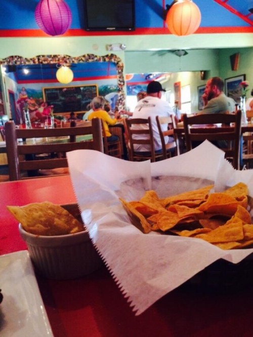 A basket of tortilla chips on a table, with a restaurant interior and patrons in the background. Colorful lanterns hang above.