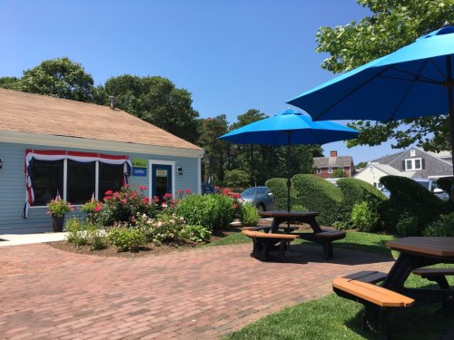 A sunny outdoor scene featuring a quaint building with colorful flowers, picnic tables, and blue umbrellas.