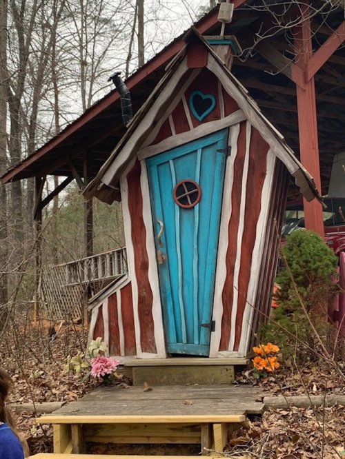 A whimsical, colorful tiny house with a blue door and striped red walls, surrounded by trees and flowers.
