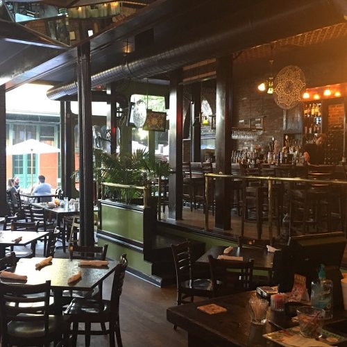 A cozy restaurant interior with wooden tables, a bar, and large windows, featuring plants and warm lighting.