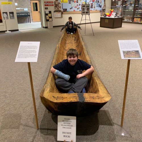 Two boys sit in a large wooden canoe exhibit in a museum, with signs instructing not to rock the canoe.