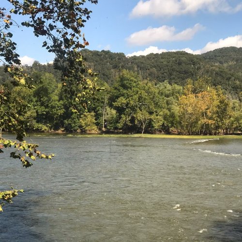 A serene river flows through a lush green landscape, surrounded by trees and distant hills under a blue sky.