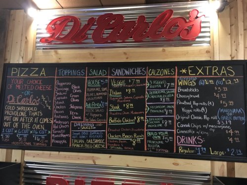 Menu board from Di Carlo's featuring pizza, toppings, salads, sandwiches, calzones, wings, and drinks with prices.