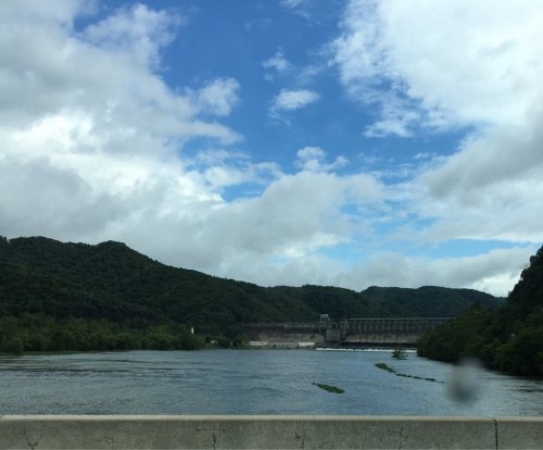 A dam stands against a backdrop of green hills and a blue sky with scattered clouds, reflecting in the water below.