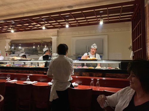 A sushi chef prepares food behind a counter while a server and a diner observe in a cozy restaurant setting.