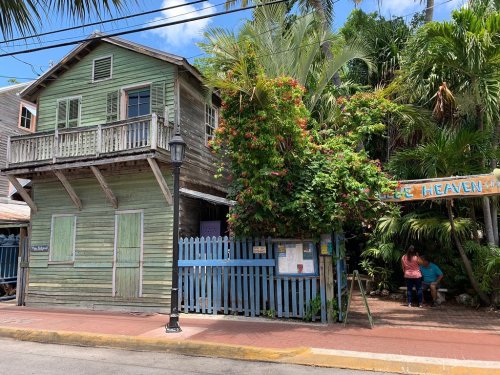 A colorful, weathered wooden house surrounded by lush greenery and a blue picket fence, with a sign reading "Ice Heaven."