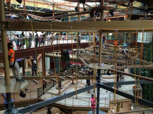 A multi-level indoor ropes course with participants navigating obstacles, surrounded by onlookers in a spacious venue.