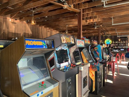 A row of vintage arcade machines, including Frogger and others, in a rustic bar setting with wooden beams.
