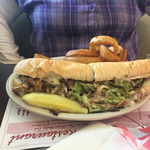 A large sandwich filled with grilled meat, peppers, and onions, served with a pickle and curly fries on the side.