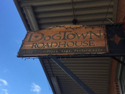 Sign for DogTown Roadhouse featuring wood-fired pizza, taps, and performances, set against a blue sky.