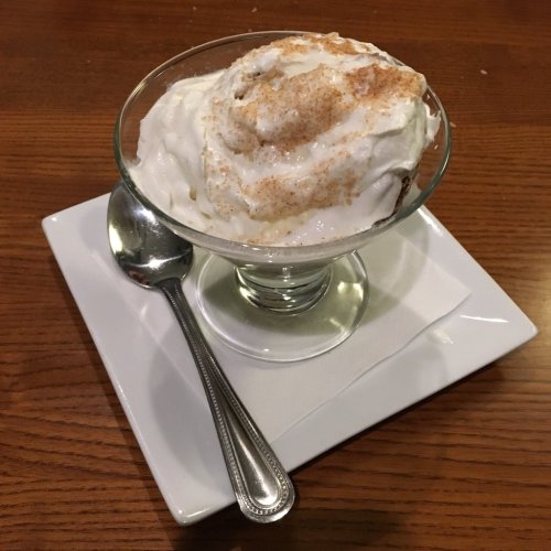 A glass dessert dish filled with whipped cream, topped with a sprinkle of cinnamon, served on a white plate with a spoon.