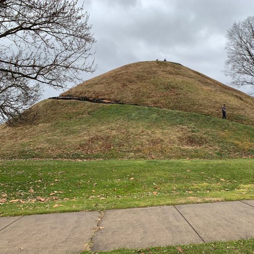 A grassy mound under a cloudy sky, with a person standing near its base and two figures on top.