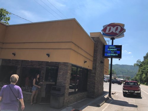 A Dairy Queen restaurant with a digital sign showing open hours, set against a clear blue sky and mountains in the background.
