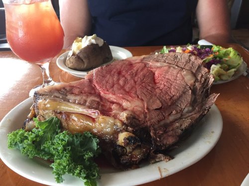 A large serving of prime rib with sides of baked potato, salad, and a drink on a wooden table.