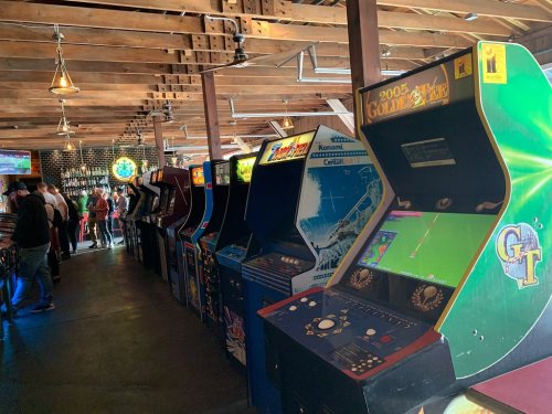 A row of vintage arcade machines in a lively bar with wooden beams and patrons in the background.