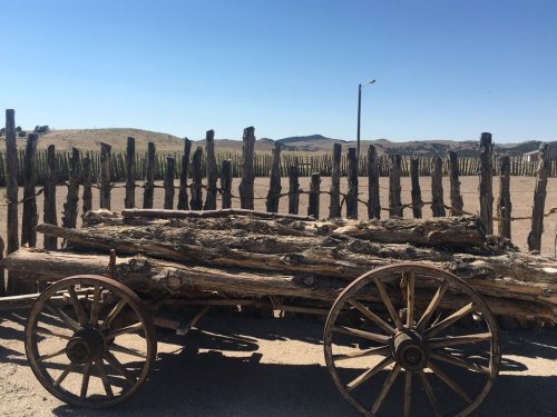 A rustic wooden cart with large wheels, set against a backdrop of a wooden fence and rolling hills under a clear blue sky.