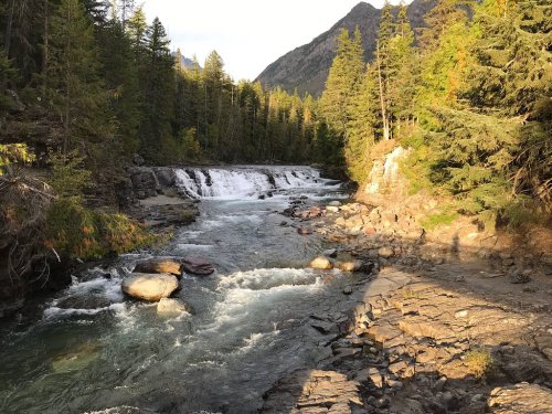 A serene river flows through a forested landscape, with a waterfall and rocky banks under soft sunlight.