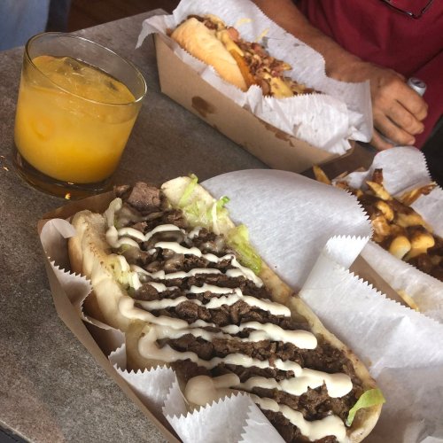 A close-up of a loaded sandwich with beef and toppings, accompanied by a drink and side dishes in the background.