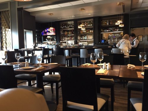 A modern restaurant interior featuring a bar, tables set for dining, and shelves stocked with drinks.
