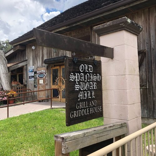 Sign for "Old Spanish Sugar Mill Grill and Griddle House" outside a rustic building with greenery and flowers.