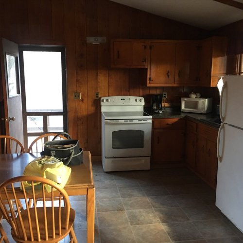 A cozy kitchen with wooden cabinets, an oven, microwave, and a dining table with chairs. Natural light from a window.