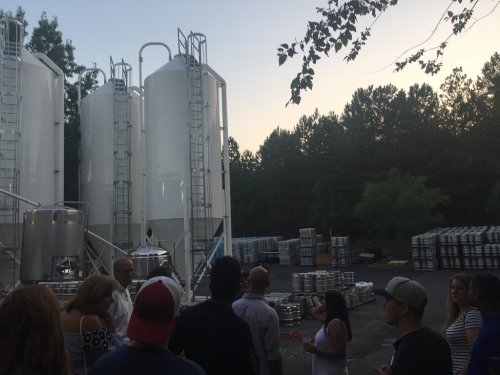 A group of people gathers near large silos and brewing equipment at a brewery during sunset.