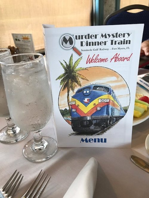 Menu for the Murder Mystery Dinner Train featuring a train illustration and tropical scenery, with drinks and fruit on the table.