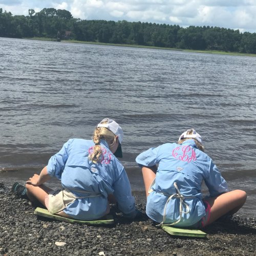 Two children in blue shirts and hats sit by a river, playing with the water on a sunny day.