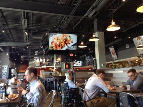 A busy restaurant interior with diners at tables, a large screen displaying food, and modern decor.
