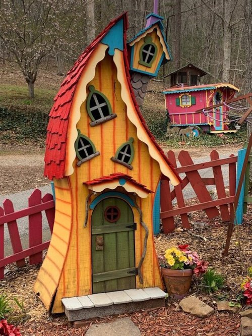 A whimsical, colorful miniature house with a slanted roof, surrounded by a wooden fence and vibrant flowers.