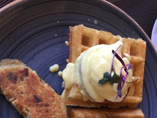 A plate featuring a waffle topped with poached egg and hollandaise sauce, alongside a slice of savory sausage.