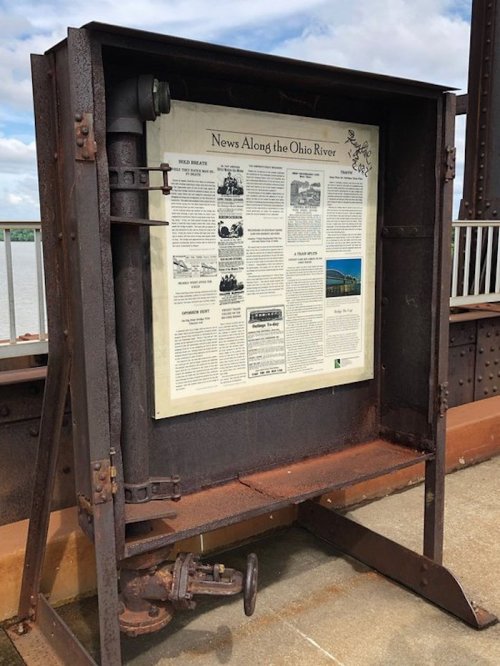 A weathered display board on a bridge features news articles about the Ohio River, surrounded by rusted metal.
