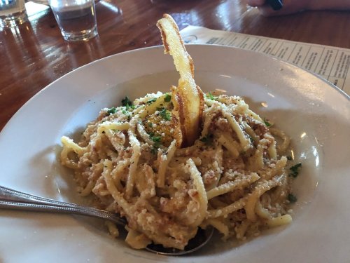 A plate of creamy pasta topped with a crispy bacon strip and garnished with herbs, served in a rustic setting.