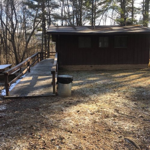 A wooden cabin surrounded by trees, with a ramp leading to the entrance and a trash can nearby on a frosty ground.