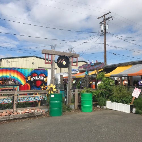 Colorful outdoor market scene with sculptures, basketball hoops, and vibrant murals under a cloudy sky.