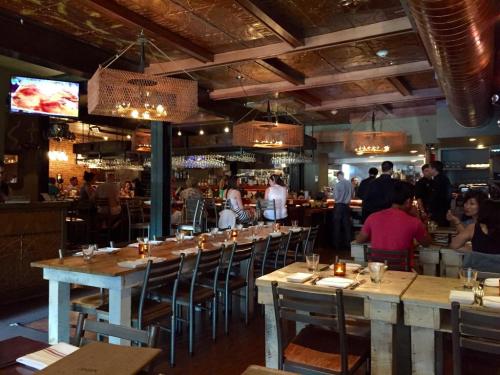 A bustling restaurant interior with wooden tables, warm lighting, and a bar area, featuring diners and staff.