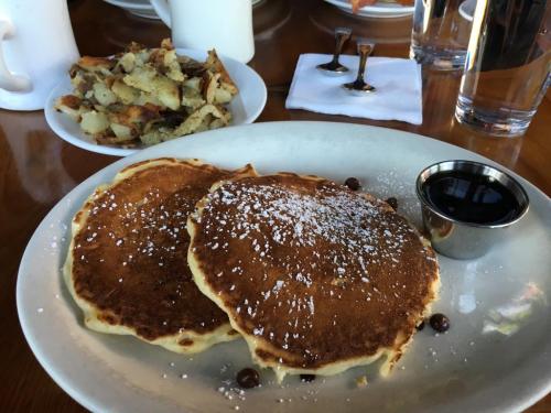Two fluffy pancakes dusted with powdered sugar, served with a side of crispy potatoes and syrup in a small cup.